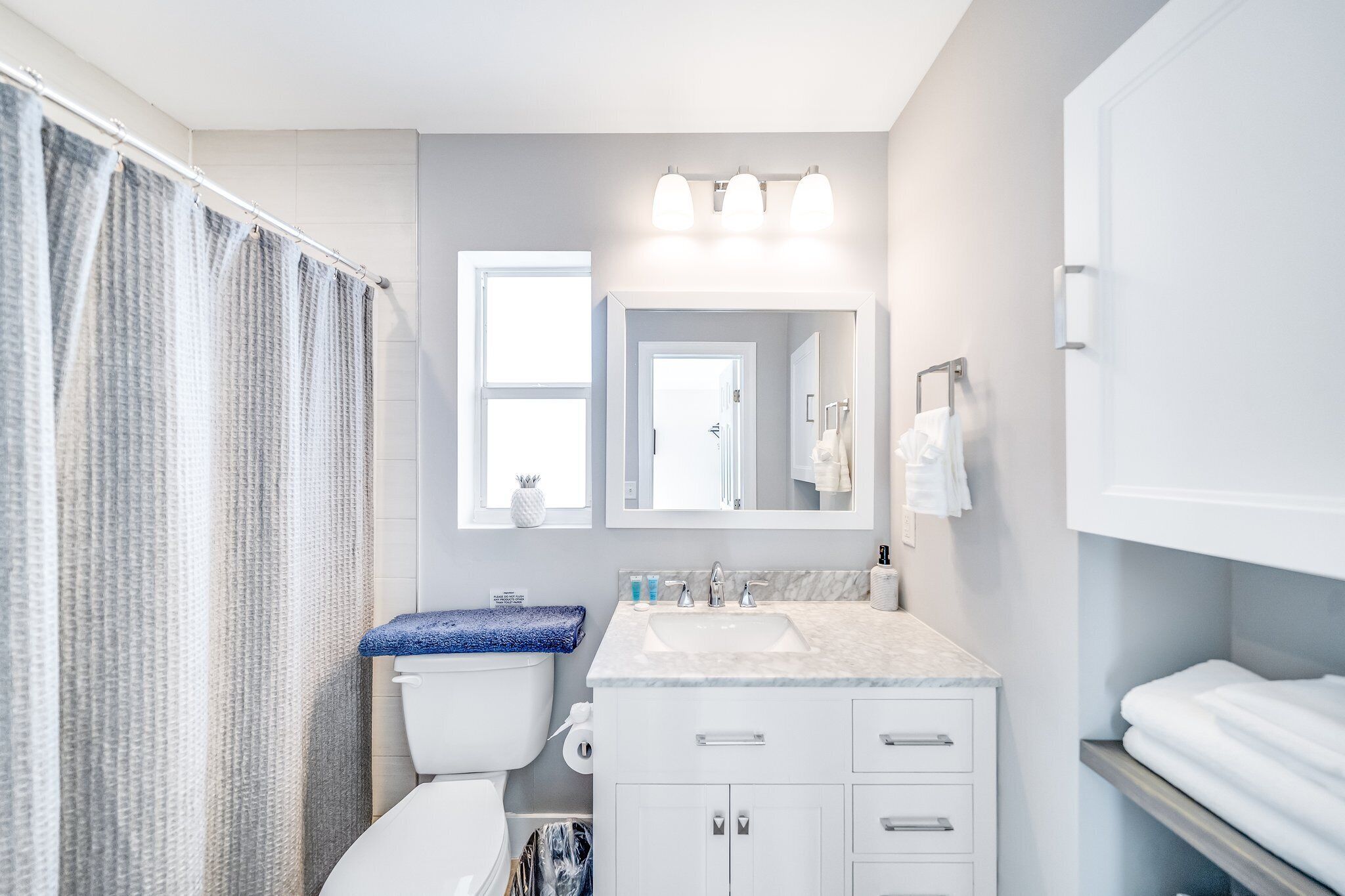 Un baño moderno con inodoro blanco, lavamanos con encimera de mármol y un espejo grande sobre el lavamanos.