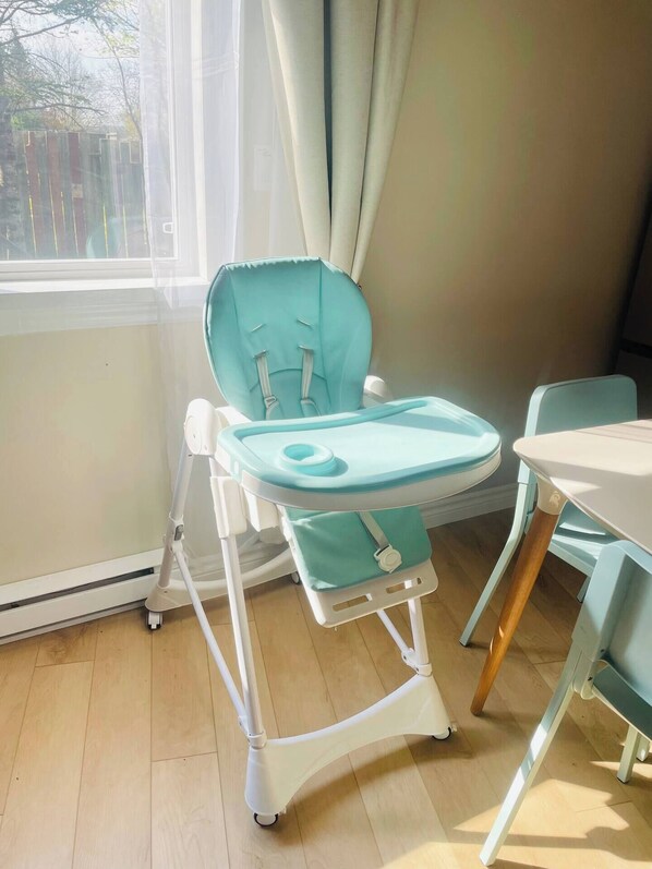 A high chair with a tray, positioned near a window with curtains, in a room with wooden flooring and a table. - private suite (the skylight) (Lower Sackville)