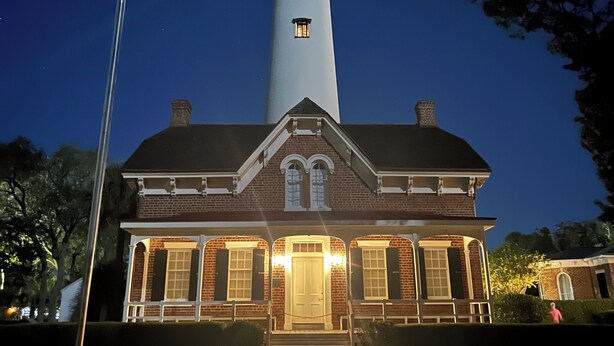 A lighthouse with a brick house in front, a brick pathway, and a clear night sky.