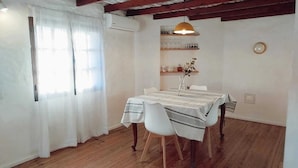 Ein Essbereich mit einem Tisch für vier Personen, weißen Stühlen, einer hängenden Pendelleuchte und einem an der Wand befestigten Regal mit Dekorationen.