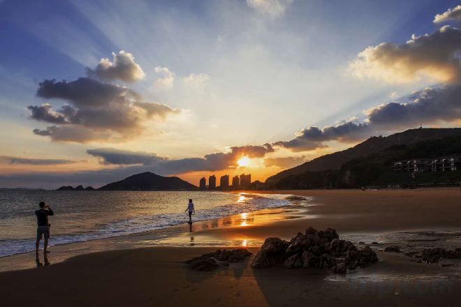 A beach scene with a sunset, a person taking a photo, and a distant cityscape.
