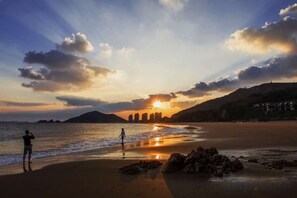 Eine Szene am Strand mit Sonnenuntergang, einer Person, die ein Foto macht, und einer weiter entfernten Skyline.