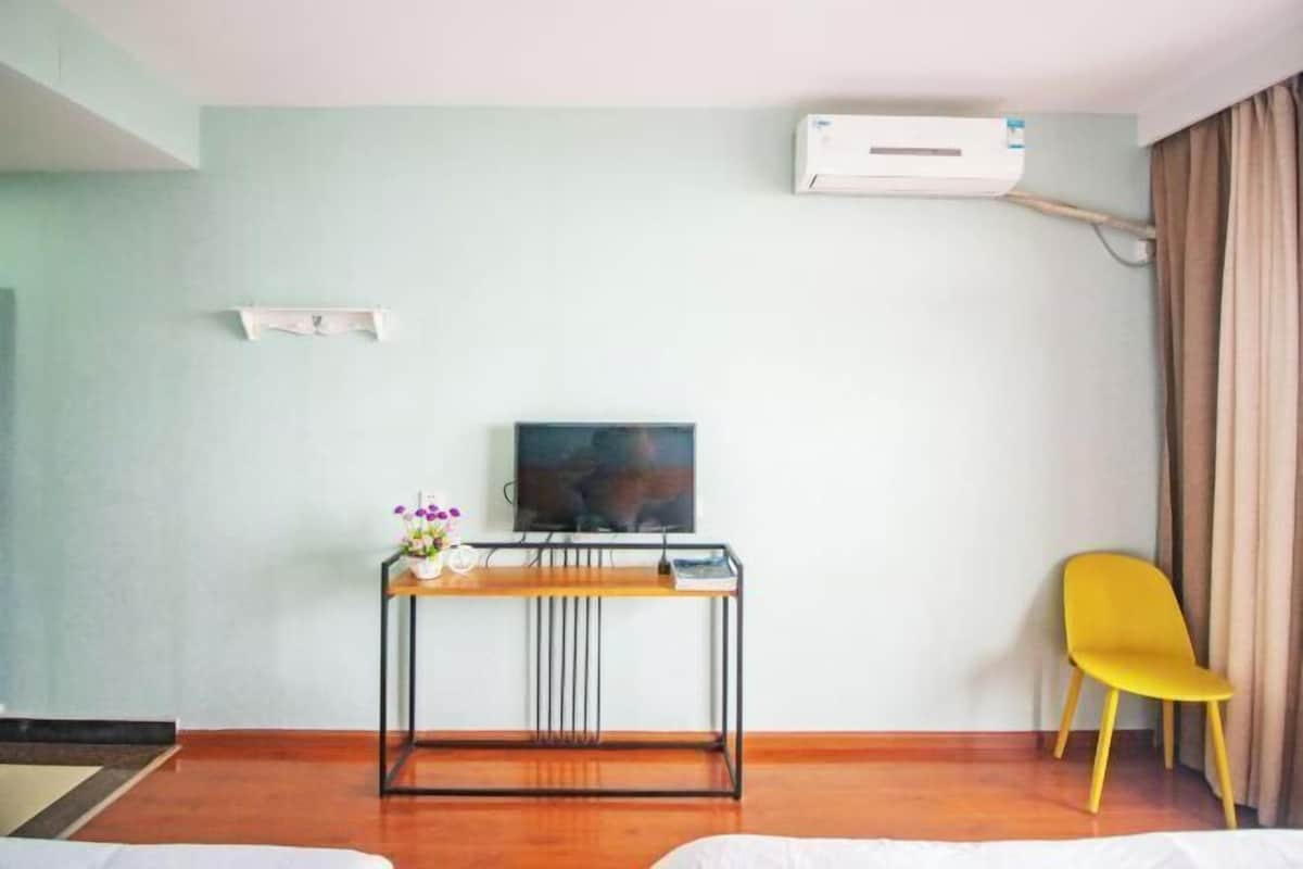 A hotel room with a bed, a desk with a TV, a yellow chair, and an air conditioning unit.