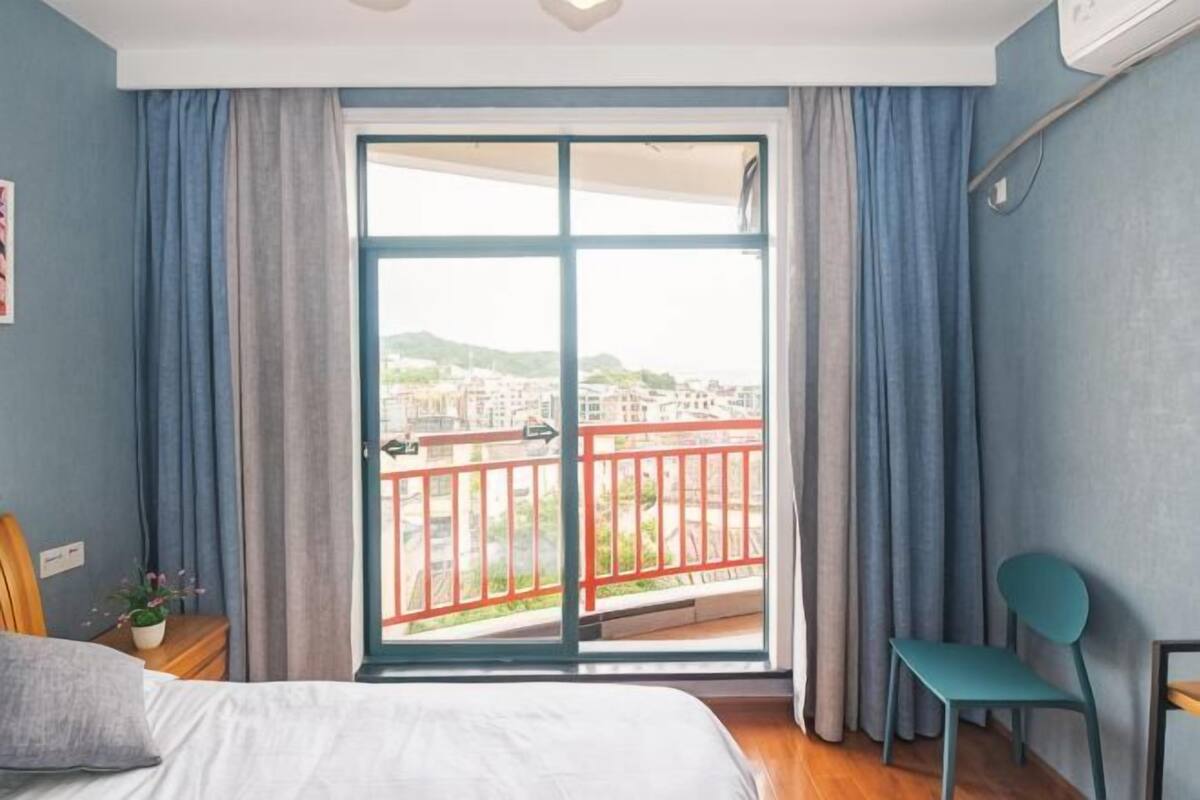 Un dormitorio con un amplio ventanal corredera a un balcón, una cama, una silla y una mesita.