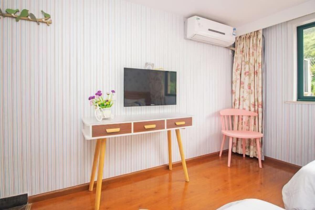 A room with a wooden floor, a white desk with drawers, a pink chair, a flat-screen TV, and a window with curtains.