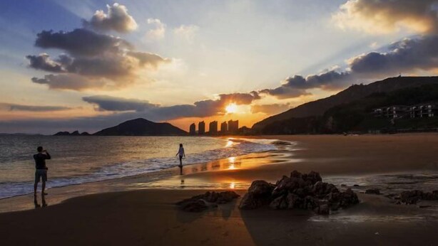 A beach scene with a sunset, rocky shoreline, and distant buildings. - Lizhou Mountain Villa (Shengsi)