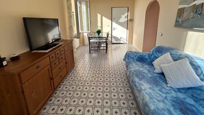 Ein Zimmer mit Fernseher auf einer Holdecke, ein blaues Sofa mit gemustertem Bezug, ein Fliesenboden, ein Esstisch mit Stühlen und ein Bild an der Wand.
