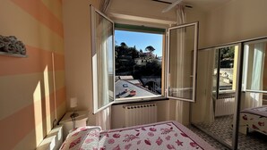 Ein Schlafzimmer mit großem Fenster, einem Bett mit floralem Überwurf, einem Schminktisch und einem Spiegel.