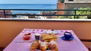 Ein Tisch mit Croissants, einer Tasse Kaffee und einem Joghurtbecher auf einem Balkon mit Blick aufs Meer.