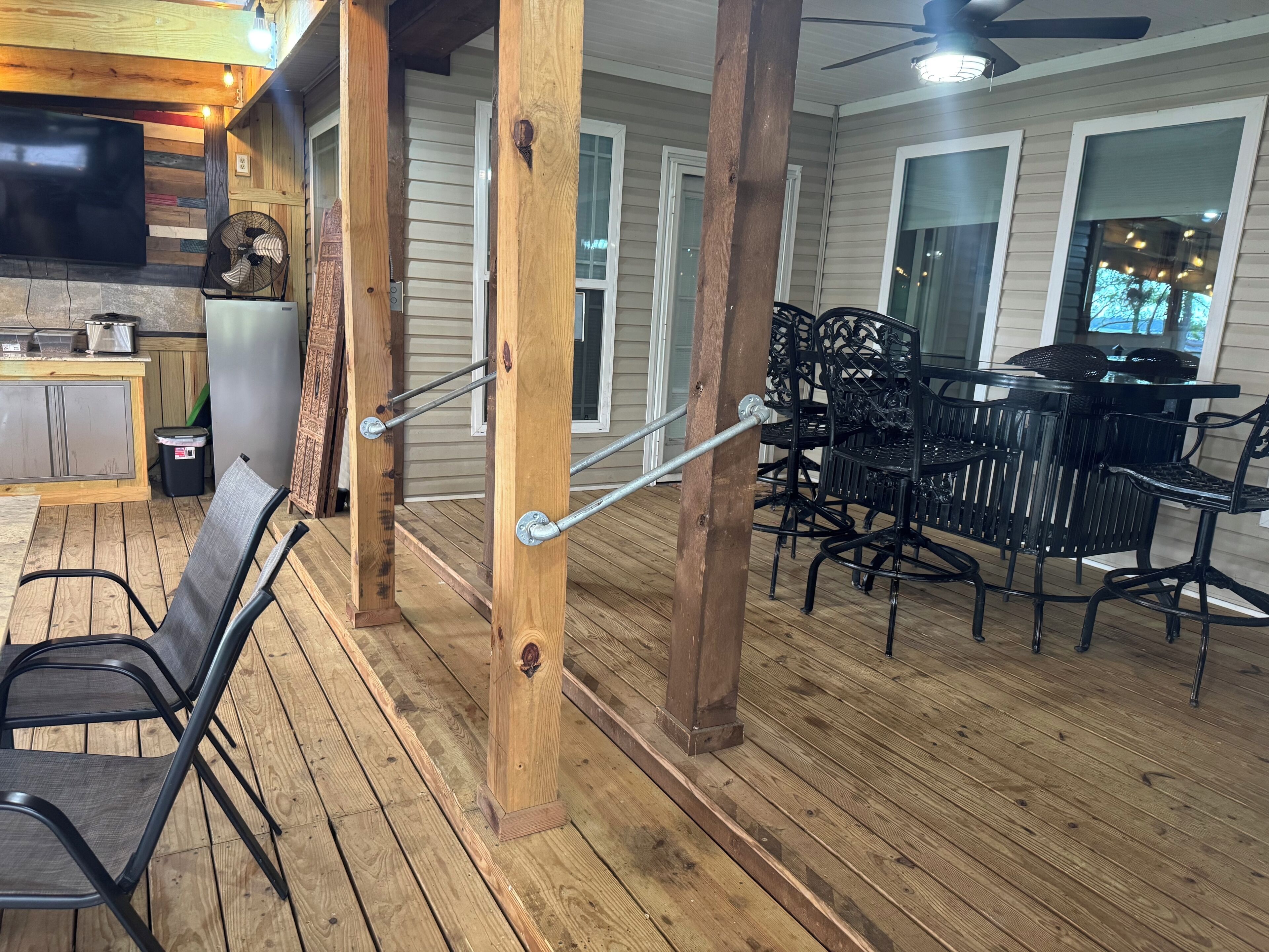 Un espacio al aire libre cubierto con piso de madera, una mesa de comedor con sillas, un ventilador de techo y un televisor montado en la pared.