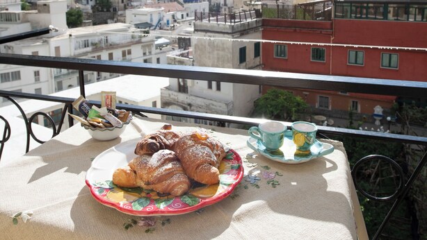 A balcony with a table set for breakfast, overlooking a coastal town with buildings and a hill.