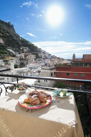 A balcony with a table set for breakfast, overlooking a coastal town with buildings and a hill.