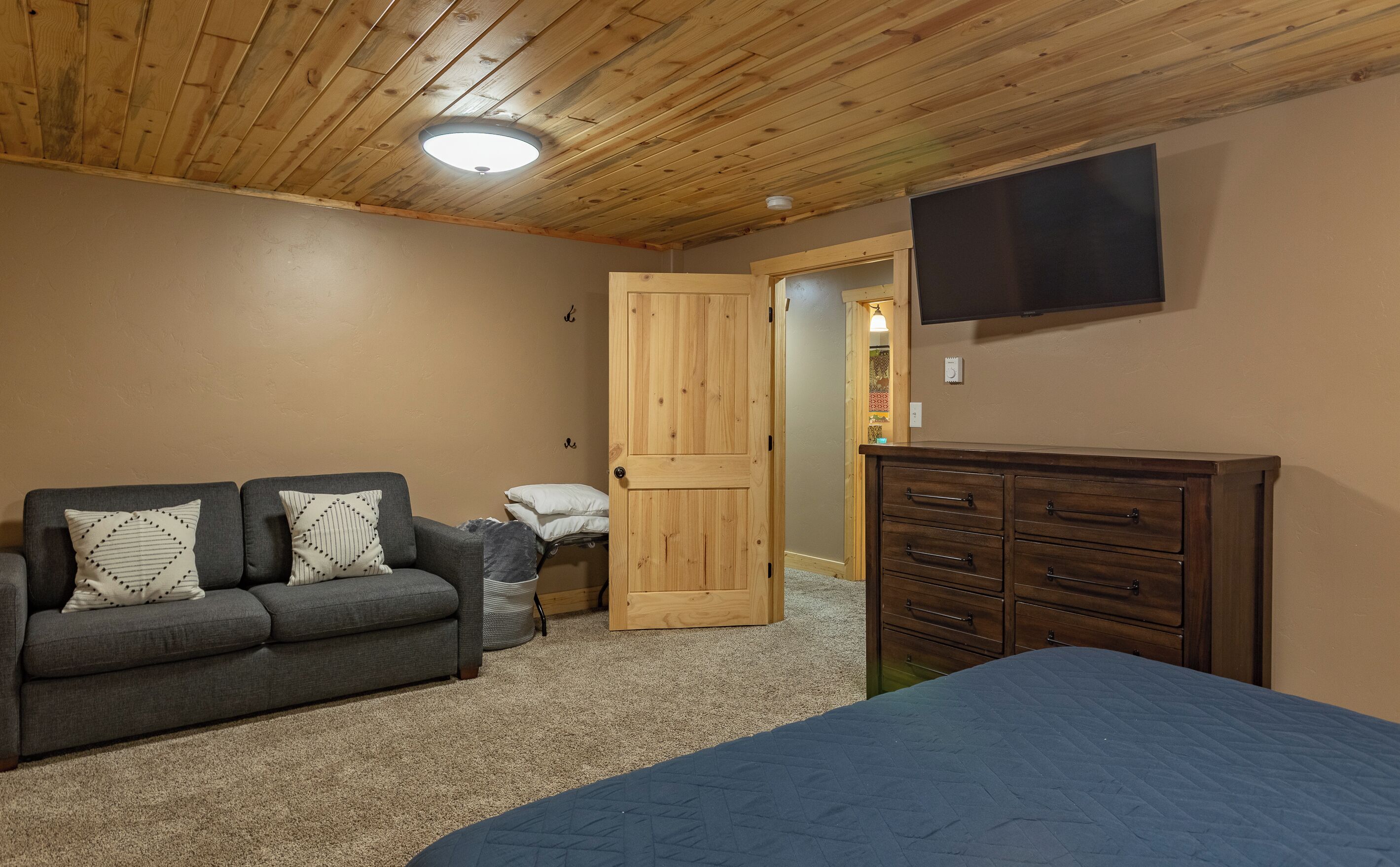 Una camera da letto con soffitto in legno, un divano grigio con cuscini decorati, un comò in legno e un letto con copripiumino blu.