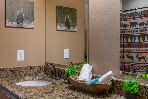 A bathroom with a granite countertop, a wooden bowl sink, a mirror, and bear-themed artwork on the wall. - Black Bear Lodge (Island Park)