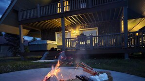 Una chimenea exterior con leña y malvaviscos, rodeada por una terraza de madera y una casa al fondo.