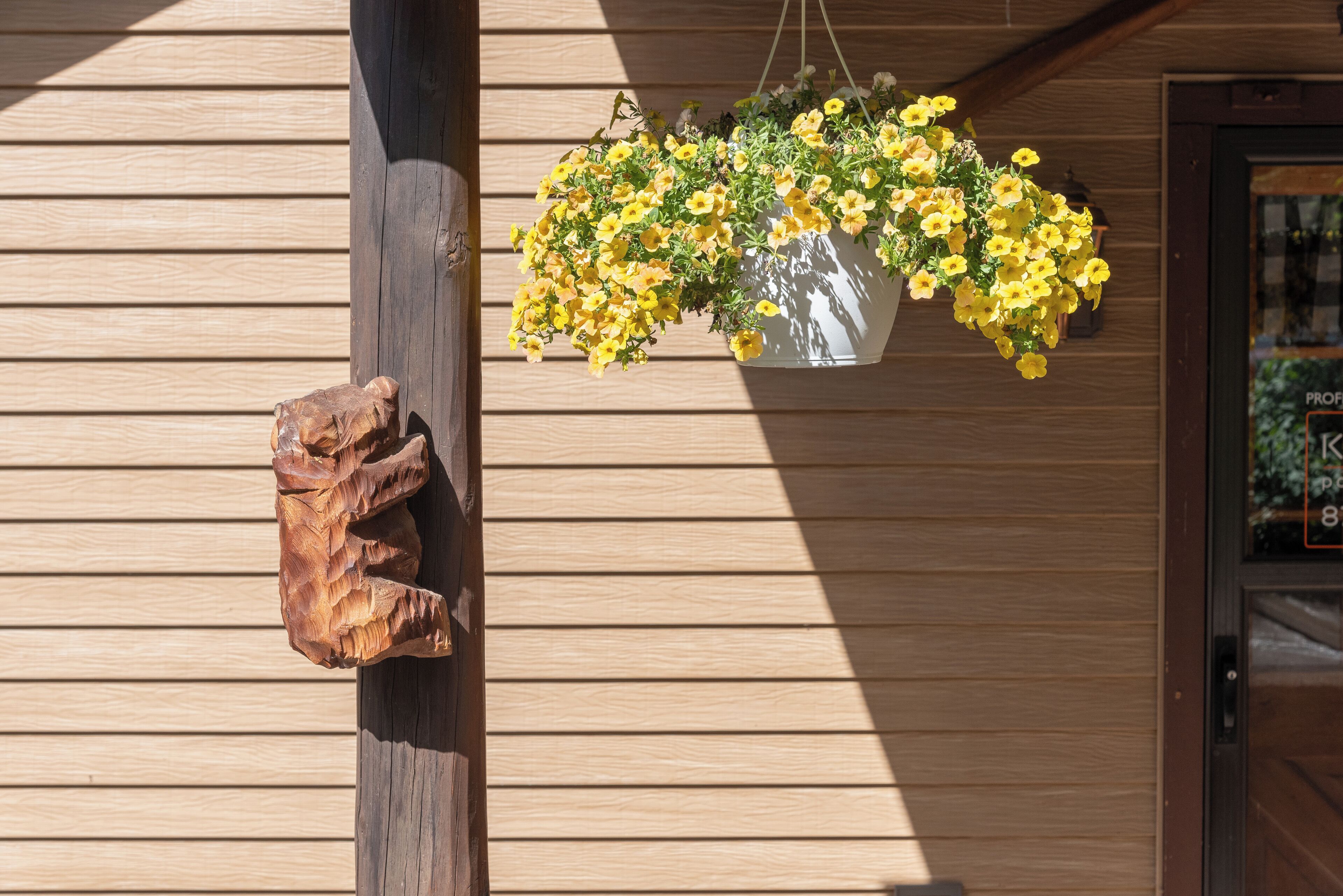 A wooden wall with horizontal planks, a hanging flower pot with yellow flowers, and a wooden sculpture.