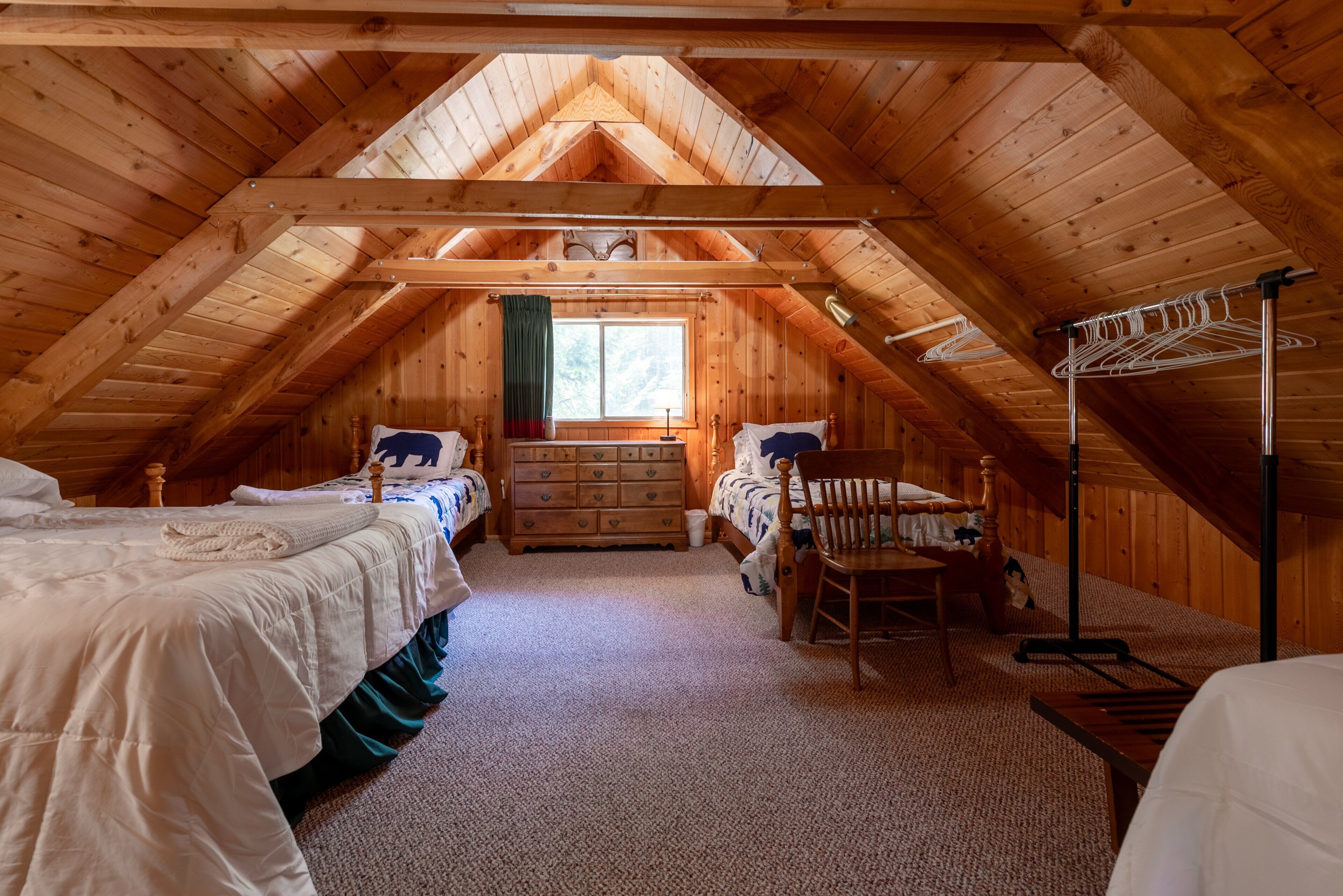 A cozy attic bedroom with wooden beams, two beds, a dresser, and a window with a view.