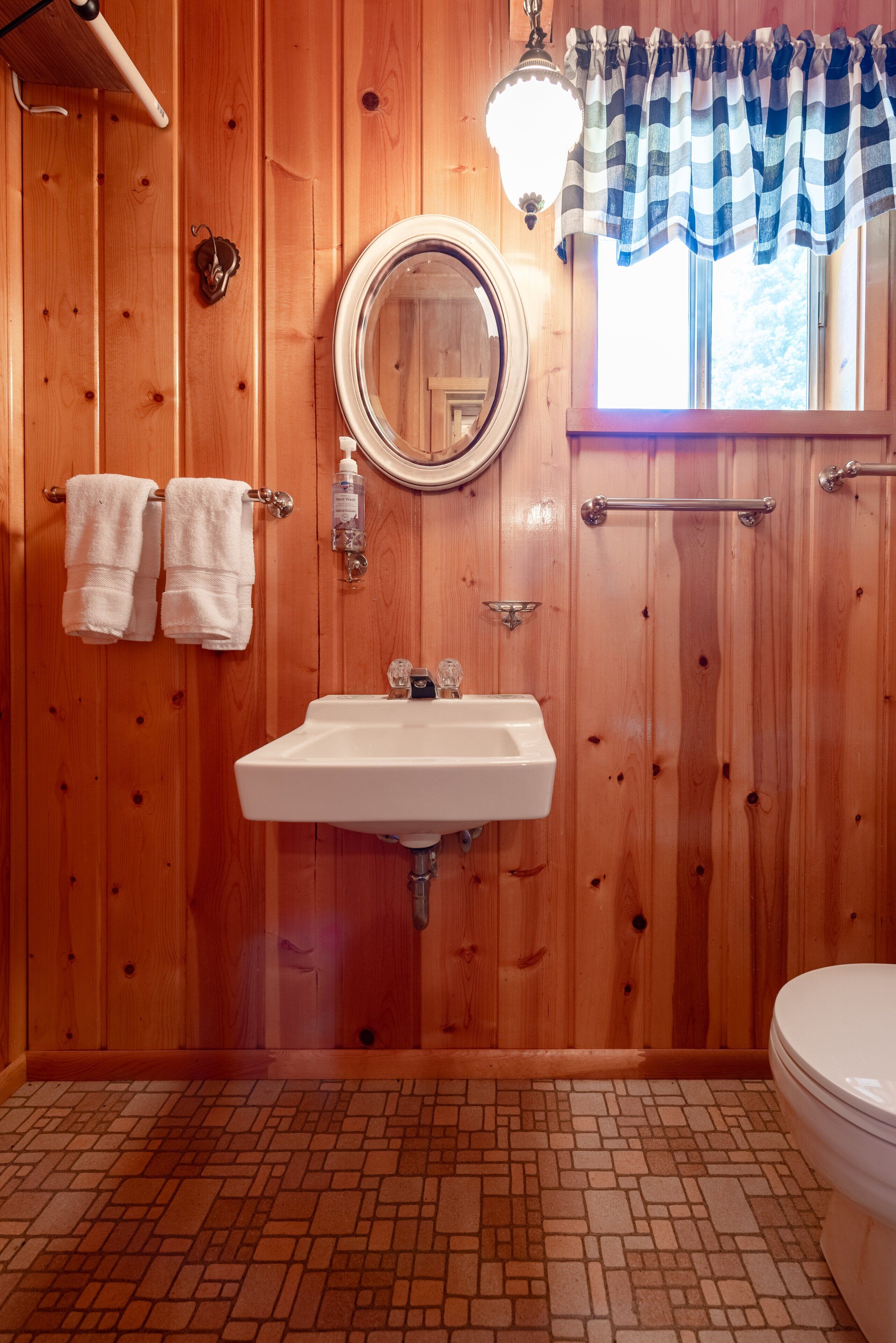 A wooden-paneled bathroom with a white sink, a round mirror, and a window with a striped valance.