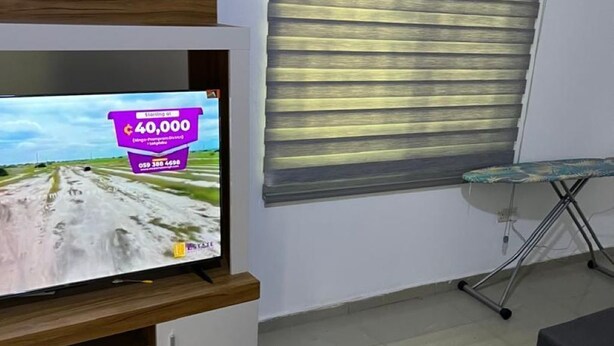 Una sala de estar con un televisor de pantalla plana montado en un mueble de madera, una ventana con persianas, una tabla de planchar de vidrio y una mesa de centro de madera.