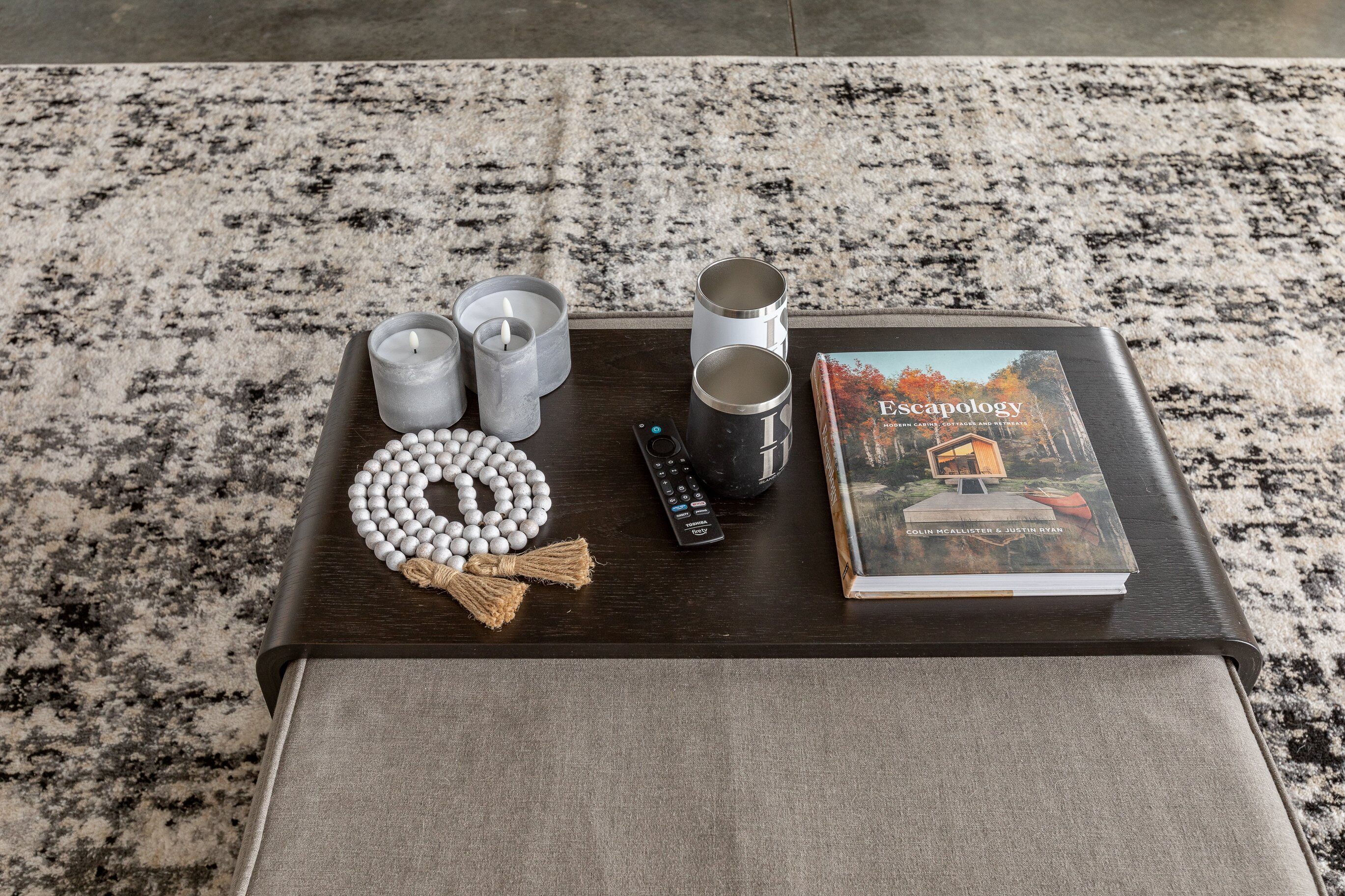 Una mesa de madera oscura con un libro titulado 'Escapology', un control remoto, velas y un collar de cuentas de oración sobre una alfombra estampada.