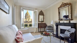 A living room with a sofa, armchairs, a small table with fruit, a fireplace, and a view of a building through a window. - Charming Apartment w/ Iconic Views of Les Invalides (Paris)