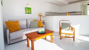Ein Wohnzimmer mit einer Couch, einem Holztisch, einem Korbstuhl, einer Lampe und einer Küche im Hintergrund.