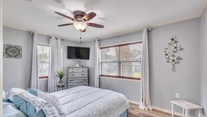 Een slaapkamer met een bed, een plafondventilator, een televisie, een dressoir en een raam met jaloezieën.