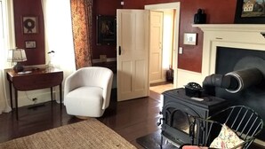 Een gezellige woonkamer met een open haard, een witte fauteuil, een bank en een rustiek plafond.