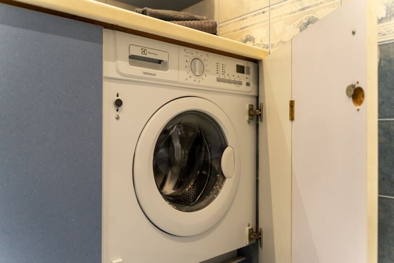Un lave-linge à chargement frontal, doté d’un panneau de commande, situé dans une salle de bain dont les murs sont recouverts de carreaux.