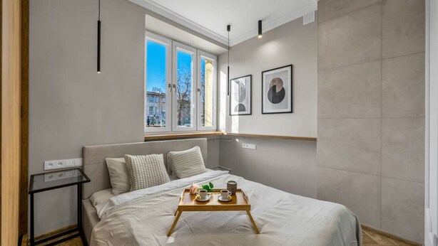Un dormitorio moderno con una cama grande, una mesita de noche pequeña, una ventana con vistas a edificios y cuadros enmarcados en la pared.