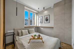 A modern bedroom with a large bed, a small bedside table, a window with a view of buildings, and framed pictures on the wall.