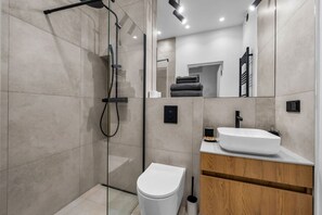 Un baño moderno con ducha envuelta en vidrio, inodoro blanco y tocador de madera con lavabo blanco.