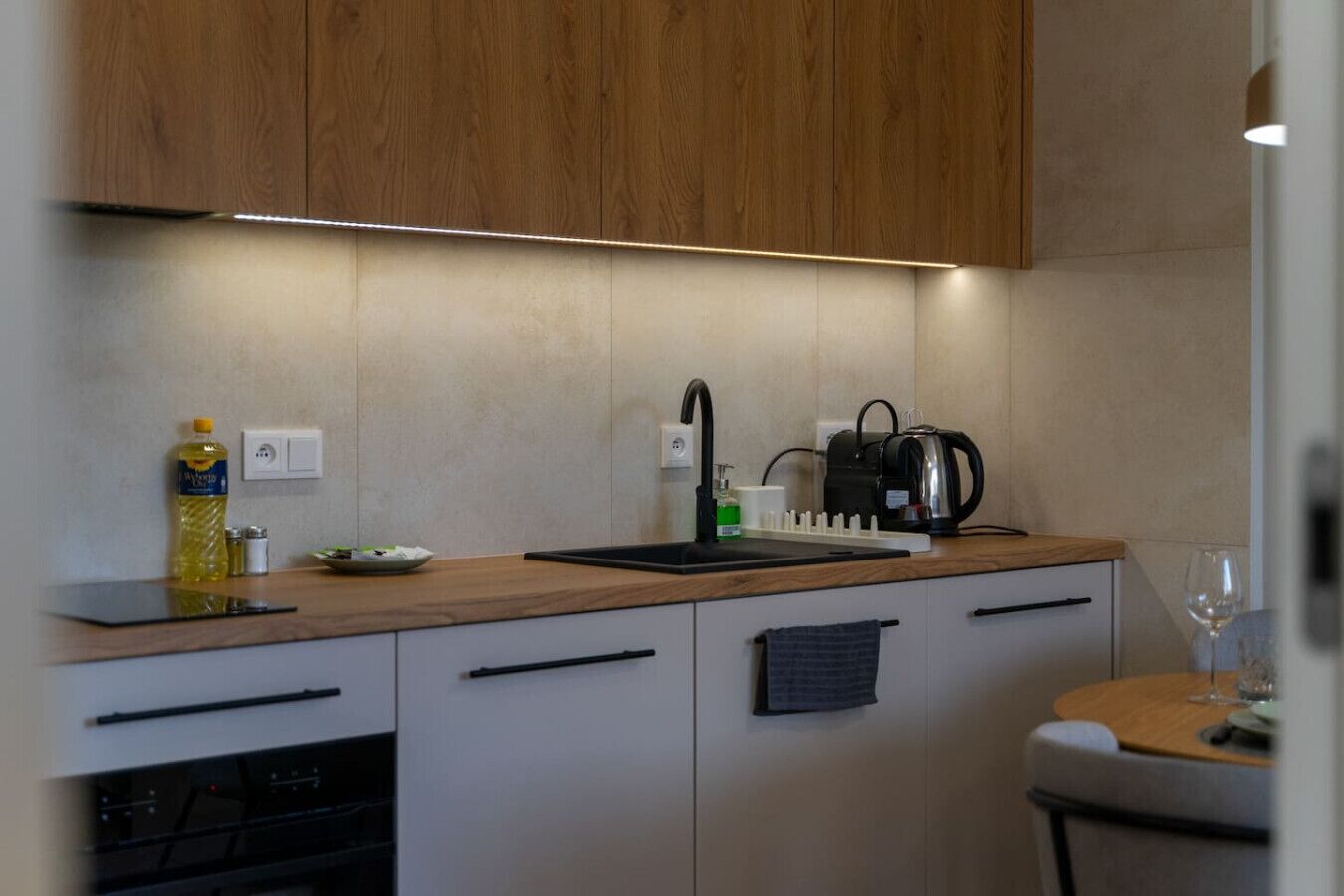 Een moderne keuken met houten kastjes, een zwart aanrecht, een spoelbak, een waterkoker en een wijnglas op een klein tafeltje.
