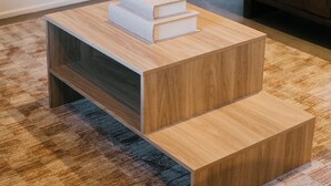 Una pequeña mesa de madera con una pila de libros y un objeto blanco encima, sobre una alfombra estampada en una habitación con un mueble de madera oscura y un televisor al fondo.
