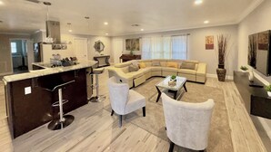 A modern living room with a curved sofa, a glass coffee table, and a kitchen area with a marble countertop and bar stools.