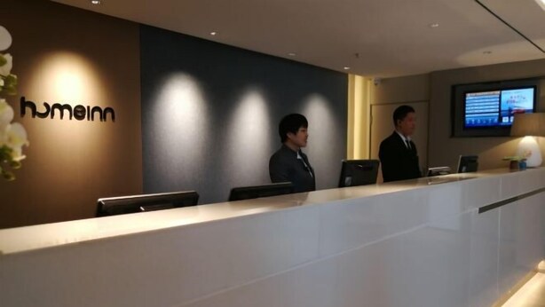 A hotel reception area with a reception desk, two staff members, and a digital screen displaying information.