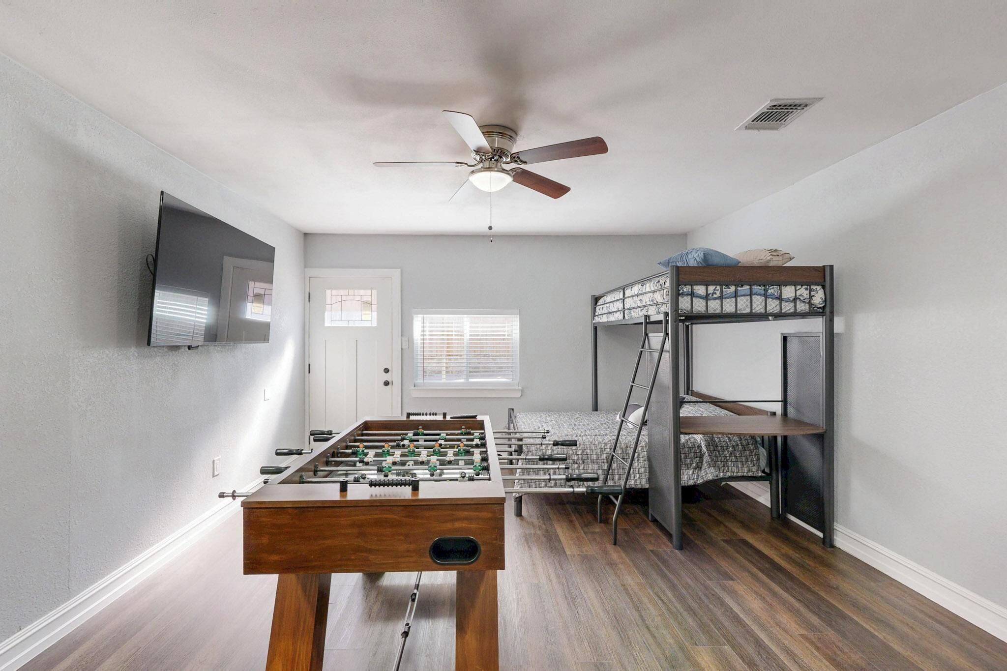 A room with a foosball table, bunk beds, a ceiling fan, a TV mounted on the wall, and a window with blinds.
