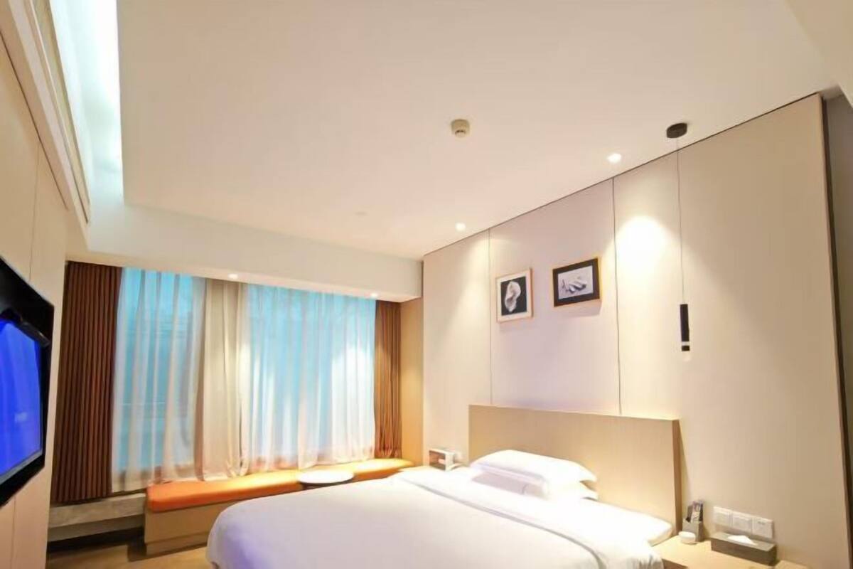 A hotel room with a bed, a TV, a bedside table, a nightstand, a window with curtains, and a wall-mounted picture.