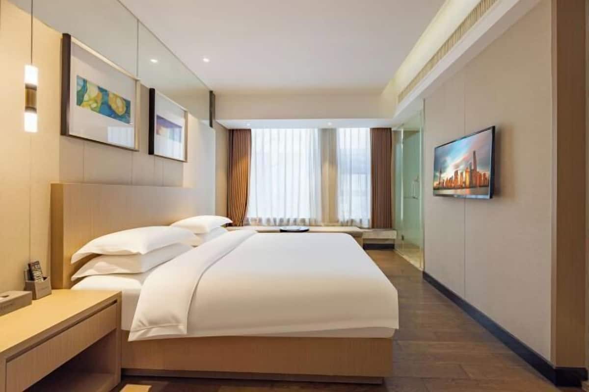 A hotel room with a large bed, a bedside table, a TV mounted on the wall, and a window with curtains.