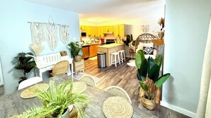 A modern kitchen with yellow cabinets, a dining area with a wooden table and chairs, and a living area with a wicker sofa and large green plants. - Boho theme, Ground floor patio with green space and creek view. 2 min to gondola (Panorama)