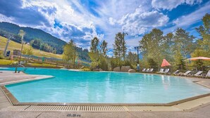A swimming pool with lounge chairs and umbrellas, surrounded by trees and a mountainous backdrop. - Boho theme, Ground floor patio with green space and creek view. 2 min to gondola (Panorama)