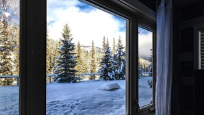 A window with a view of a snowy landscape, including snow-covered trees and a snow-covered ground. - Boho theme, Ground floor patio with green space and creek view. 2 min to gondola (Panorama)