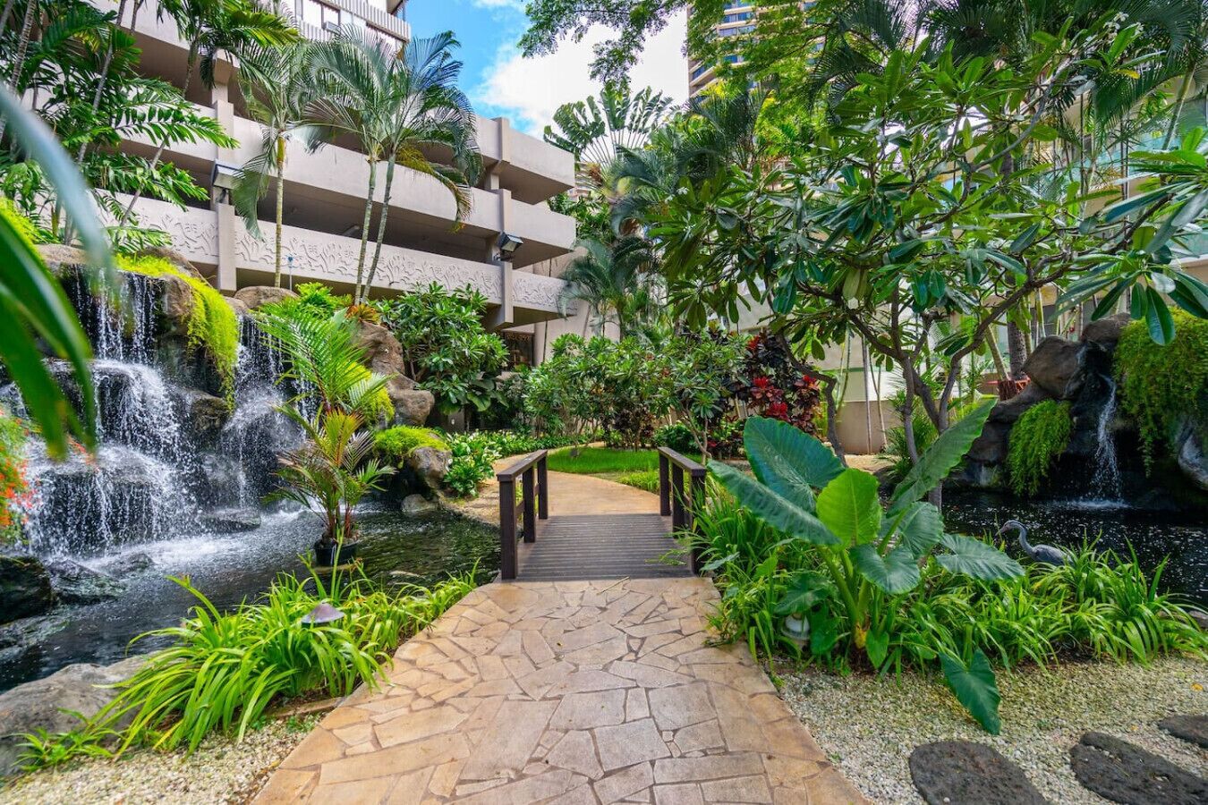 A tropical garden with a stone pathway leading to a wooden bridge, surrounded by lush greenery and a cascading waterfall.