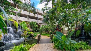 Un jardin tropical avec un sentier de pierre menant à un pont en bois, entouré d’une végétation luxuriante et d’une cascade.