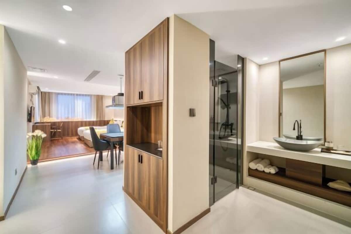 A modern hotel room with a wooden wardrobe, a glass shower enclosure, and a bathroom with a large mirror and sink.