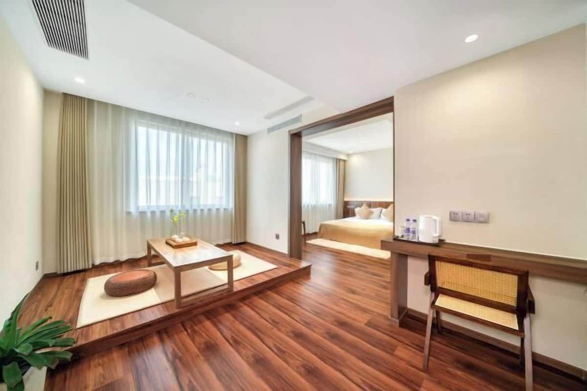 A modern hotel room with a wooden floor, a bed with white bedding, a small table with a cushion, a chair, and a bedside table with a lamp.