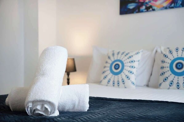 A neatly made bed with a rolled white towel, a lamp, and patterned pillows on a dark blue bedspread.