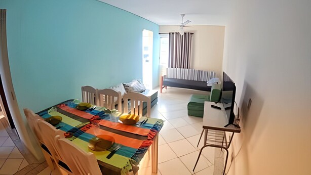 A dining area with a table set for four, a sofa, a TV, and a ceiling fan in the background. - Conforto Praia de Leste (Pontal do Parana)