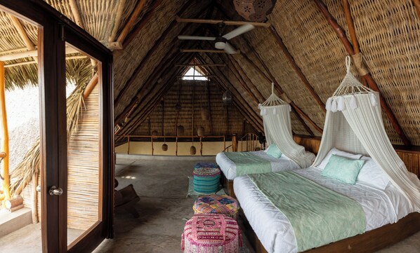 A thatched-roof room with a bed, a hanging chair, and a view of palm trees.