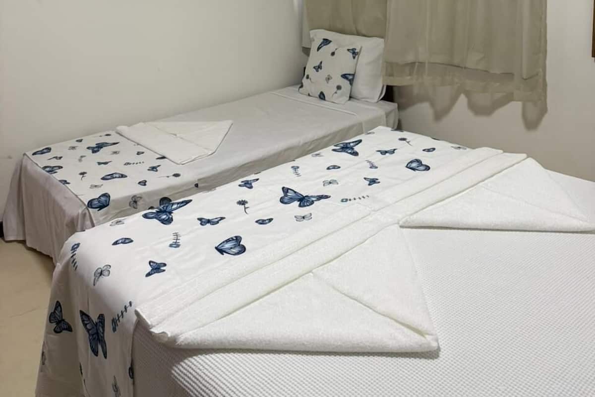 A room with two single beds, a flat-screen TV mounted on the wall, and a window with sheer curtains.